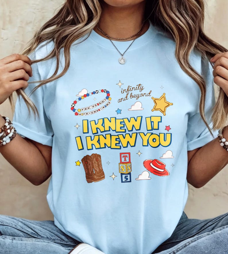 May include: A light blue t-shirt featuring a graphic print with the text "I KNEW IT I KNEW YOU", "infinity and beyond", and "YOU'VE GOT A FRIEND IN ME". The design includes illustrations of a beaded bracelet, a star, cowboy boots, blocks, and a hat.