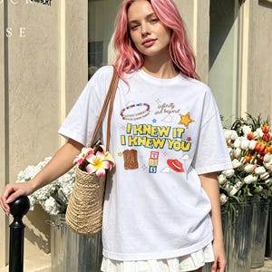 May include: A white short-sleeved t-shirt featuring a graphic print with the text I KNEW IT I KNEW YOU, infinity and beyond, a beaded bracelet, a yellow star, a brown cowboy boot, alphabet blocks, and a red hat with a woven straw bag.