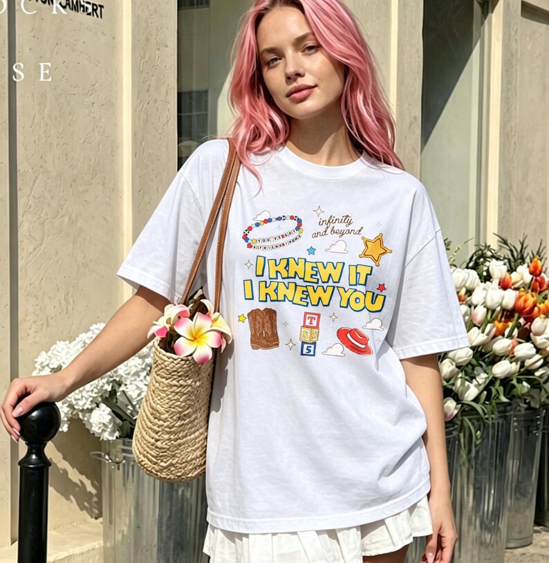 May include: A white short-sleeved t-shirt featuring a graphic print with the text I KNEW IT I KNEW YOU, infinity and beyond, a beaded bracelet, a yellow star, a brown cowboy boot, alphabet blocks, and a red hat with a woven straw bag.