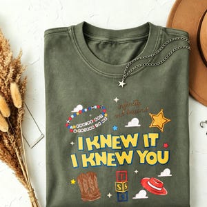 May include: An olive green crew neck t-shirt featuring a graphic print with the text "I KNEW IT I KNEW YOU" in yellow, "YOU'VE GOT A FRIEND IN ME" on a beaded bracelet, and "infinity and beyond" in script, alongside cowboy boots, blocks, and a hat.