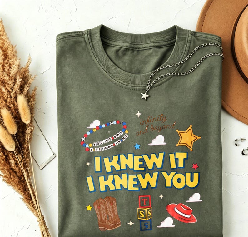 May include: An olive green crew neck t-shirt featuring a graphic print with the text "I KNEW IT I KNEW YOU" in yellow, "YOU'VE GOT A FRIEND IN ME" on a beaded bracelet, and "infinity and beyond" in script, alongside cowboy boots, blocks, and a hat.
