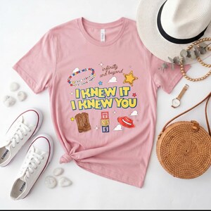 May include: A light pink short-sleeve t-shirt featuring a graphic print with the text "I KNEW IT I KNEW YOU" and "infinity and beyond," accompanied by illustrations of a beaded bracelet, a yellow star, cowboy boots, alphabet blocks, and a red hat.