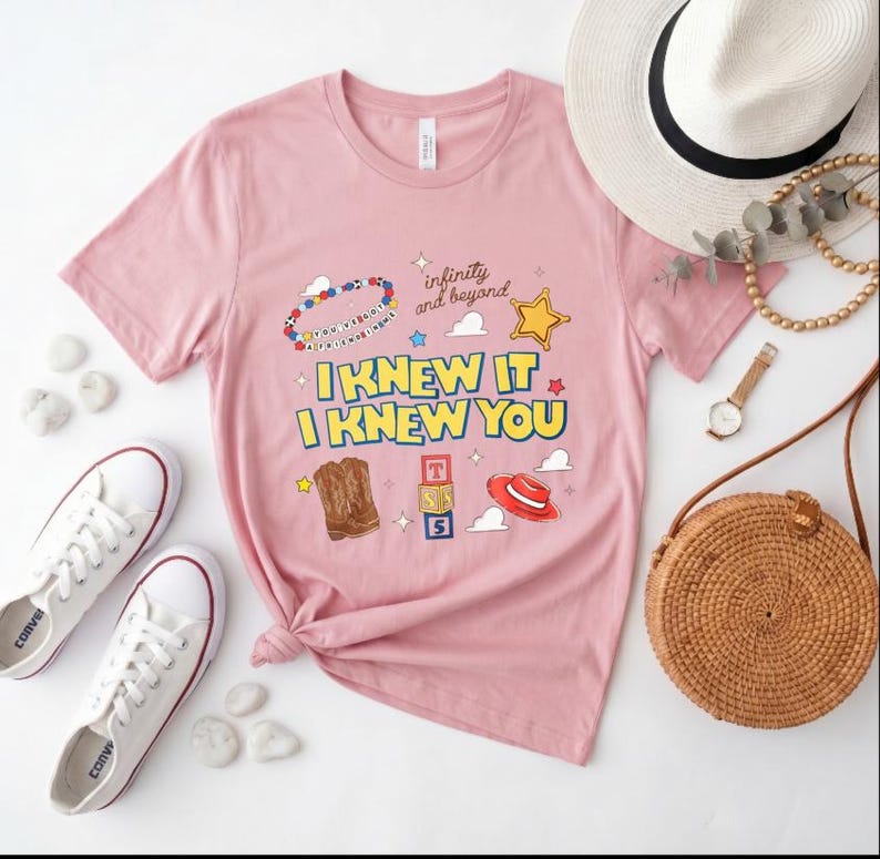 May include: A light pink short-sleeve t-shirt featuring a graphic print with the text "I KNEW IT I KNEW YOU" and "infinity and beyond," accompanied by illustrations of a beaded bracelet, a yellow star, cowboy boots, alphabet blocks, and a red hat.