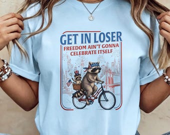Maglietta girocollo azzurra con stampa divertente di un procione patriottico "Get In Loser", citazione sulla libertà e skyline di New York con bicicletta, perfetta per il 4 luglio.