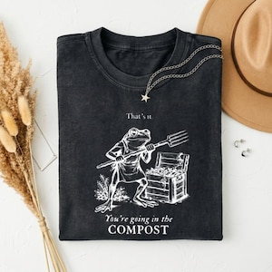 Könnte beinhalten: Ein schwarzes T-Shirt mit Rundhalsausschnitt und einer weißen Strichzeichnung eines Frosches, der eine Schürze trägt und eine Heugabel neben einer Holzkiste hält. Der Text lautet That's it. You're going in the COMPOST. Stoff mit verwaschener Optik.