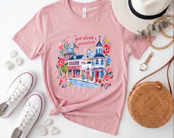 Maglietta patriottica rosa con stampa floreale e di piccola città, ideale per il 4 luglio e l'estate, con la scritta "God Bless America" e un paesaggio urbano ad acquerello.