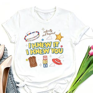 May include: A white short-sleeve t-shirt featuring a graphic print with the text "I KNEW IT I KNEW YOU" in yellow and blue. The design includes a beaded bracelet, a gold star, brown cowboy boots, alphabet blocks, and a red hat.