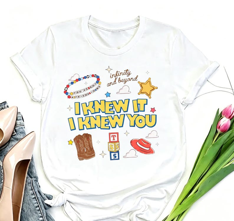 May include: A white short-sleeve t-shirt featuring a graphic print with the text "I KNEW IT I KNEW YOU" in yellow and blue. The design includes a beaded bracelet, a gold star, brown cowboy boots, alphabet blocks, and a red hat.