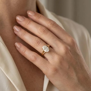 Pode incluir: Um anel de ouro com um grande diamante de corte oval. O anel tem um design de haste dividida. O diamante é cravejado com garras. O anel está no dedo de uma pessoa.