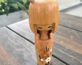 Puede incluir: Muñeca Kokeshi de madera con cabeza redondeada y cuerpo cilíndrico. La muñeca presenta una cara pintada con ojos cerrados y un diseño de kimono sencillo. El cuerpo está decorado con motivos florales y de hojas en rojo, blanco y negro.