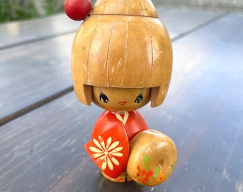 May include: A wooden Japanese Kokeshi doll with a round head and a red ball on top. The doll wears a red kimono with a white flower design and holds a brown ball with painted floral accents. The doll is a traditional Japanese craft.
