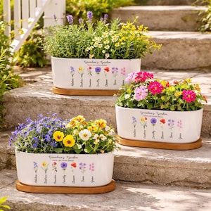 Może przedstawiać: Trzy białe ceramiczne donice wypełnione kolorowymi kwiatami i zielenią, każda na drewnianej tacy. Donice są oznaczone napisem "Grandma's Garden" z ilustracjami kwiatowymi i nazwami. Scena rozgrywa się na zewnętrznych schodach.