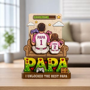 以下が含まれることがあります： ビデオゲームをする父と子のピクセル化されたデザインの木製装飾品。「PAPA」の文字が緑と黄色で表示され、その下に「I UNLOCKED THE BEST PAPA」のテキストがあります。