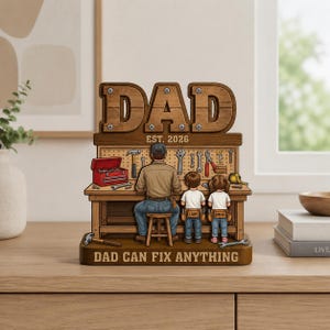 Può includere: Una statuetta in legno con la scritta "DAD" e "EST. 2026" in alto. La statuetta raffigura un uomo a un banco da lavoro con due bambini. Il testo "DAD CAN FIX ANYTHING" è alla base. Gli strumenti sono esposti sul banco da lavoro.