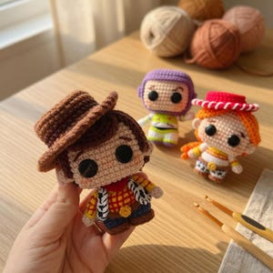 May include: Crocheted figures of Woody, Buzz Lightyear, and Jessie from Toy Story. Woody wears a brown hat and vest. Buzz has a purple hood and green and white suit. Jessie has a red hat and orange hair. Yarn balls and crochet hooks are in the background.
