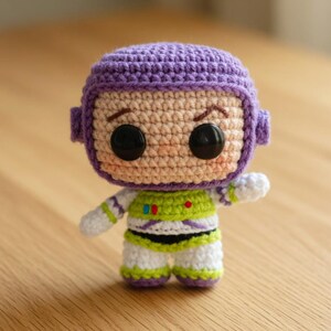 May include: A handmade crochet doll of a cartoon character. The doll features a purple helmet, tan face, and a green and white spacesuit. The doll has large black eyes and is posed with one arm raised. The doll is approximately 4 inches tall.