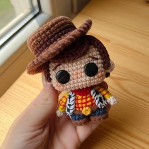 May include: Crocheted Woody doll featuring a brown cowboy hat, a red and yellow plaid shirt, blue pants, and a sheriff's badge. The doll has large black button eyes and is held in a hand. The doll is approximately 4 inches tall.