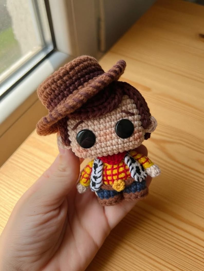 May include: Crocheted Woody doll featuring a brown cowboy hat, a red and yellow plaid shirt, blue pants, and a sheriff's badge. The doll has large black button eyes and is held in a hand. The doll is approximately 4 inches tall.