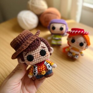 May include: Crocheted Woody doll with a brown hat, red neckerchief, and yellow shirt. Other crocheted character dolls are in the background. The dolls are handmade and appear to be part of a collection.