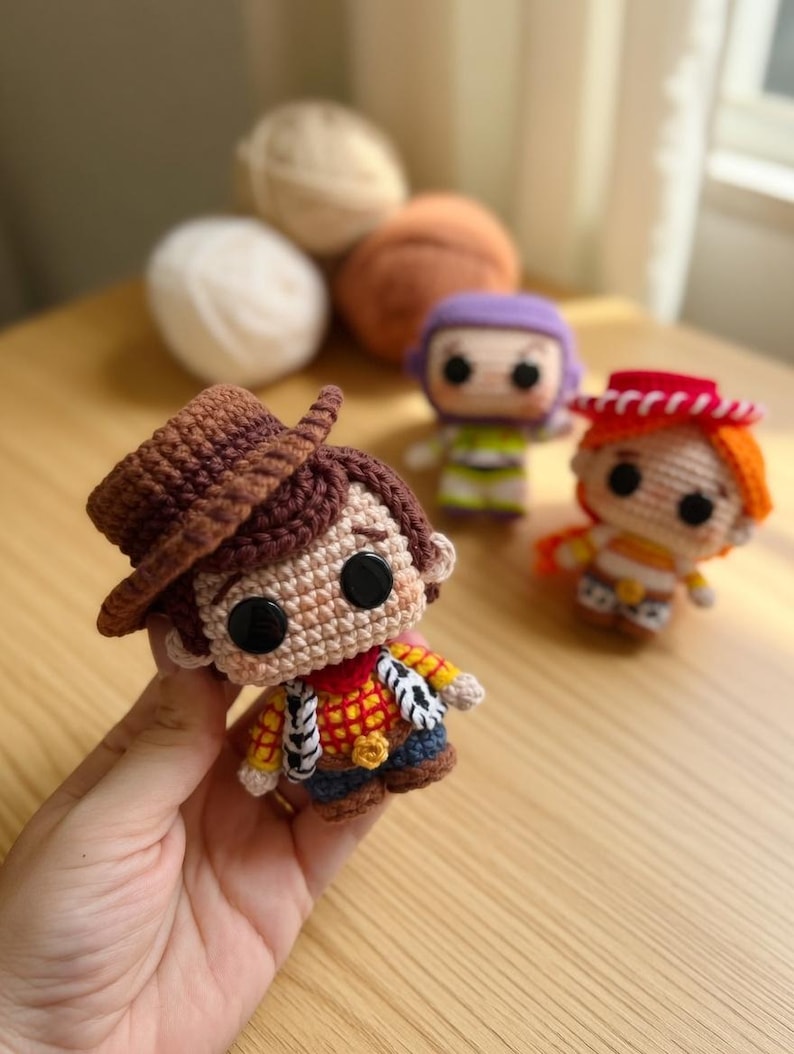 May include: Crocheted Woody doll with a brown hat, red neckerchief, and yellow shirt. Other crocheted character dolls are in the background. The dolls are handmade and appear to be part of a collection.