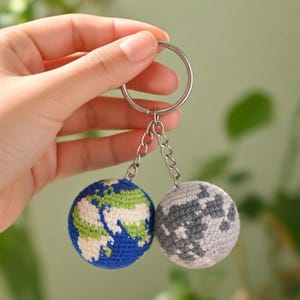 May include: A keychain featuring two crocheted spheres: one representing Earth, with blue oceans and green continents, and the other a gray and white moon. The spheres are attached to a silver keyring with small chains. The background is a soft green.