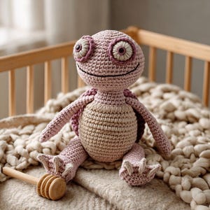 May include: A handmade crochet turtle doll in shades of pink and tan. The turtle has large, expressive eyes, a smiling mouth, and detailed feet. It is sitting on a soft, textured blanket, with a wooden honey dipper in the foreground.