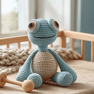 May include: A handmade crochet turtle doll in shades of blue and tan. The turtle has large, expressive eyes and a smiling mouth. The doll is sitting on a woven surface, with a wooden rattle nearby. The toy is designed for children.