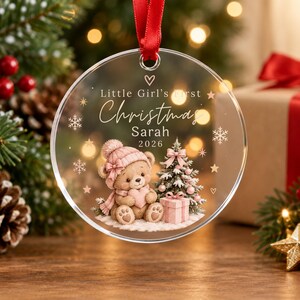 Könnte beinhalten: Ein runder, transparenter Acryl-Weihnachtsanhänger an einem roten Band. Er zeigt den Text "Little Girl's first Christmas Sarah 2026" sowie einen braunen Teddybären mit rosa Mütze und Schal, einen kleinen geschmückten Baum und ein Geschenk.