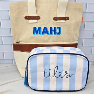 May include: A beige tote bag with cream handles and brown leather details, embroidered with the letters "MAHJ" in blue. A matching blue and white striped cosmetic bag with the word "tiles" embroidered on it.