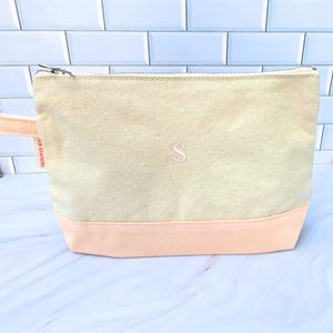 May include: A canvas cosmetic bag with a light peach base and a light beige upper section. The bag has a zipper closure and a small peach-colored handle. An embroidered pink letter "S" is centered on the front.