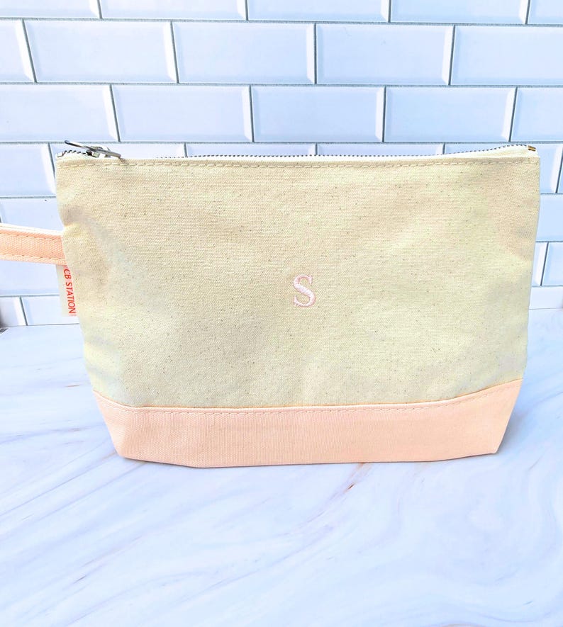 May include: A canvas cosmetic bag with a light peach base and a light beige upper section. The bag has a zipper closure and a small peach-colored handle. An embroidered pink letter "S" is centered on the front.