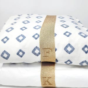 May include: Two folded white fabric items stacked vertically. The top item features a blue diamond pattern with radiating lines. A wide, textured gold band wraps around both items, with the letter F embroidered on the top and K on the bottom.