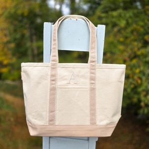 May include: A beige canvas tote bag with a light brown base, trim, and handles. The bag features a front pocket and a large embroidered letter "A". The tote is sitting on a light blue chair.