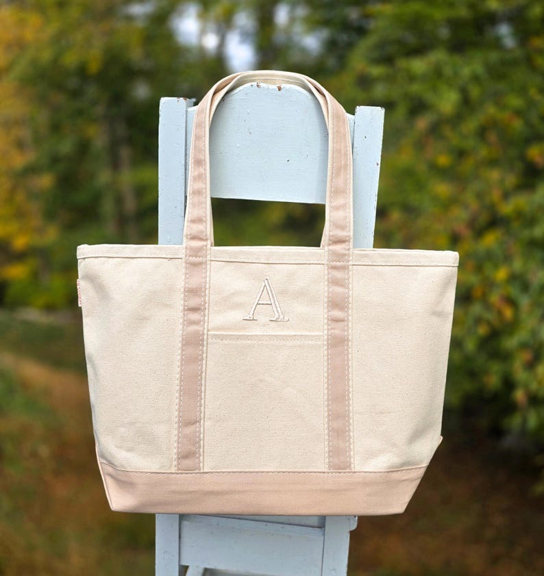 May include: A beige canvas tote bag with a light brown base, trim, and handles. The bag features a front pocket and a large embroidered letter "A". The tote is sitting on a light blue chair.