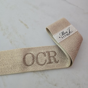 Puede incluir: Una banda el&aacute;stica de color dorado met&aacute;lico con las letras OCR bordadas en hilo oscuro. Una etiqueta de tela blanca est&aacute; unida al extremo de la banda, mostrando el texto negro Mrs. T. HANDMADE en una combinaci&oacute;n de letra cursiva y de molde.