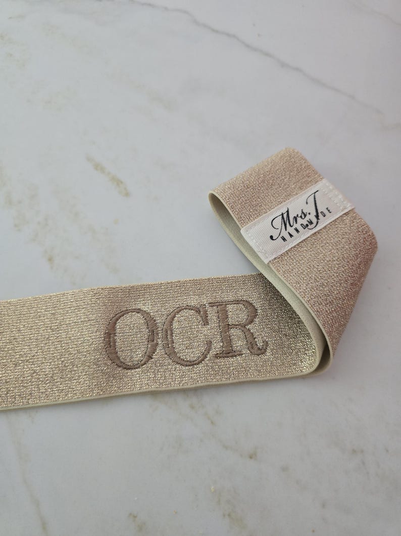 Puede incluir: Una banda el&aacute;stica de color dorado met&aacute;lico con las letras OCR bordadas en hilo oscuro. Una etiqueta de tela blanca est&aacute; unida al extremo de la banda, mostrando el texto negro Mrs. T. HANDMADE en una combinaci&oacute;n de letra cursiva y de molde.