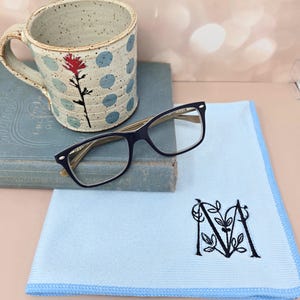 May include: A ceramic mug with blue polka dots and a red floral design, resting on a vintage book. A pair of black-framed eyeglasses and a light blue microfiber cloth with an embroidered "M" are also present.