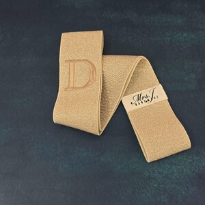 Puede incluir: Una banda el&aacute;stica de color dorado que presenta una letra D grande bordada en un extremo y una peque&ntilde;a etiqueta de tela blanca con el texto impreso en negro Mrs. T. HANDMADE unida al otro extremo del material brillante y plegado.