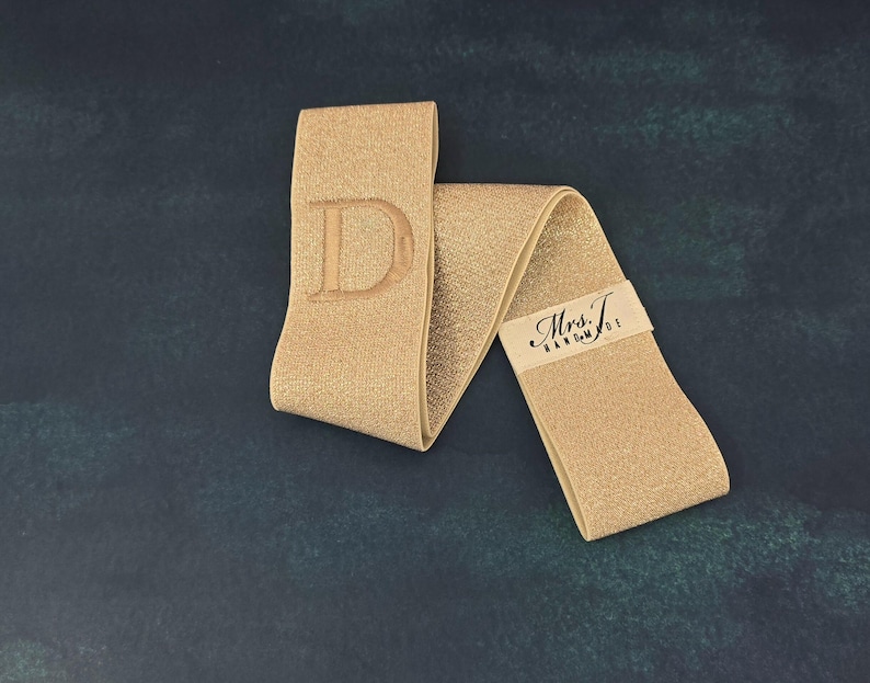 Puede incluir: Una banda el&aacute;stica de color dorado que presenta una letra D grande bordada en un extremo y una peque&ntilde;a etiqueta de tela blanca con el texto impreso en negro Mrs. T. HANDMADE unida al otro extremo del material brillante y plegado.