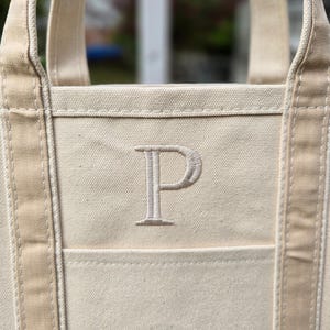 May include: Cream-colored canvas tote bag with a large embroidered letter "P" on the front. The bag has sturdy handles and a pocket. The neutral color and simple design make it a versatile accessory.