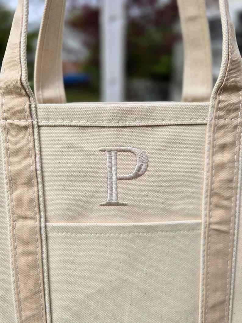 May include: Cream-colored canvas tote bag with a large embroidered letter "P" on the front. The bag has sturdy handles and a pocket. The neutral color and simple design make it a versatile accessory.