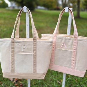 May include: Two canvas tote bags with a neutral color scheme. One bag has a light brown trim and the letter "P" embroidered on the front pocket. The other bag has a pink trim and the letter "A" embroidered on the front pocket. Both bags have long handles.