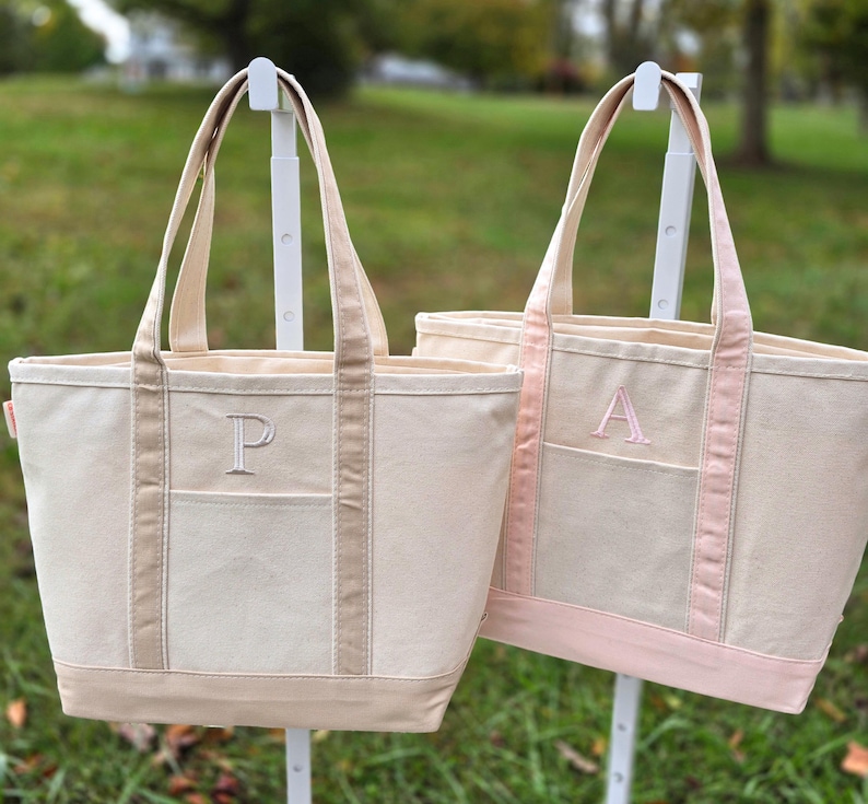 May include: Two canvas tote bags with a neutral color scheme. One bag has a light brown trim and the letter "P" embroidered on the front pocket. The other bag has a pink trim and the letter "A" embroidered on the front pocket. Both bags have long handles.