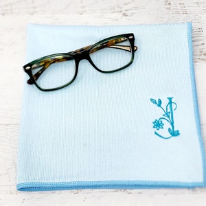 May include: A light blue microfiber cleaning cloth with a blue embroidered floral design and a pair of green eyeglasses on top.