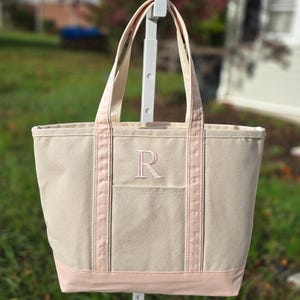 May include: A beige canvas tote bag with pink accents. The bag features a pink embroidered letter "R" on the front pocket and pink trim along the top and bottom. The tote has two beige handles.