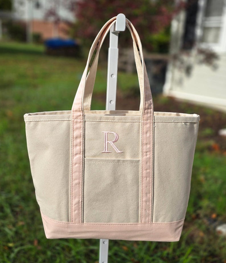 May include: A beige canvas tote bag with pink accents. The bag features a pink embroidered letter "R" on the front pocket and pink trim along the top and bottom. The tote has two beige handles.