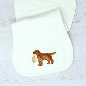 May include: Off-white baby bib with an embroidered brown dog holding a letter "B" in its mouth. The dog has a red collar. The bib is made of soft fabric and is designed for a baby.
