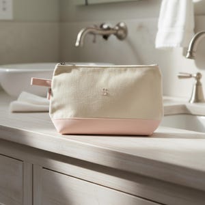 May include: A cream-colored canvas cosmetic bag with a pink base and a small pink handle. The bag has a silver zipper and a small embroidered "S" on the front. The bag is sitting on a light-colored countertop.