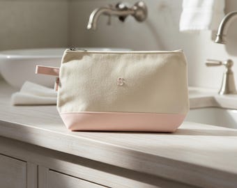 May include: A cream-colored canvas cosmetic bag with a pink base and a small pink handle. The bag has a silver zipper and a small embroidered "S" on the front. The bag is sitting on a light-colored countertop.