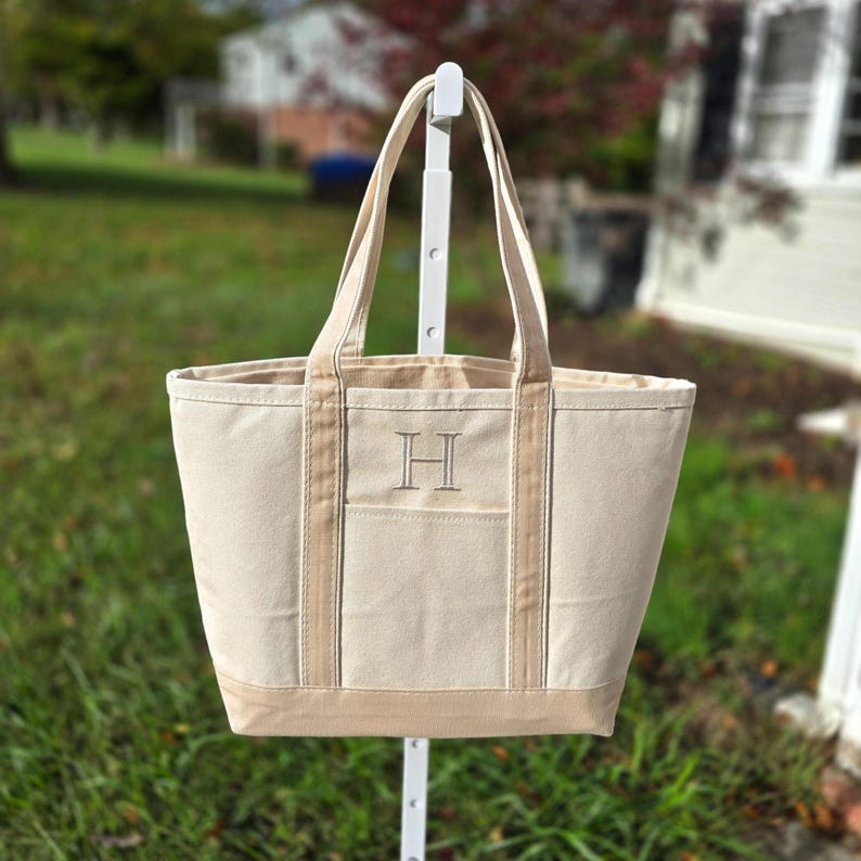 May include: A beige canvas tote bag with a light brown base, side accents, and handles. The bag features a front pocket and an embroidered letter "H". The tote is displayed outdoors, suggesting a casual, everyday accessory.